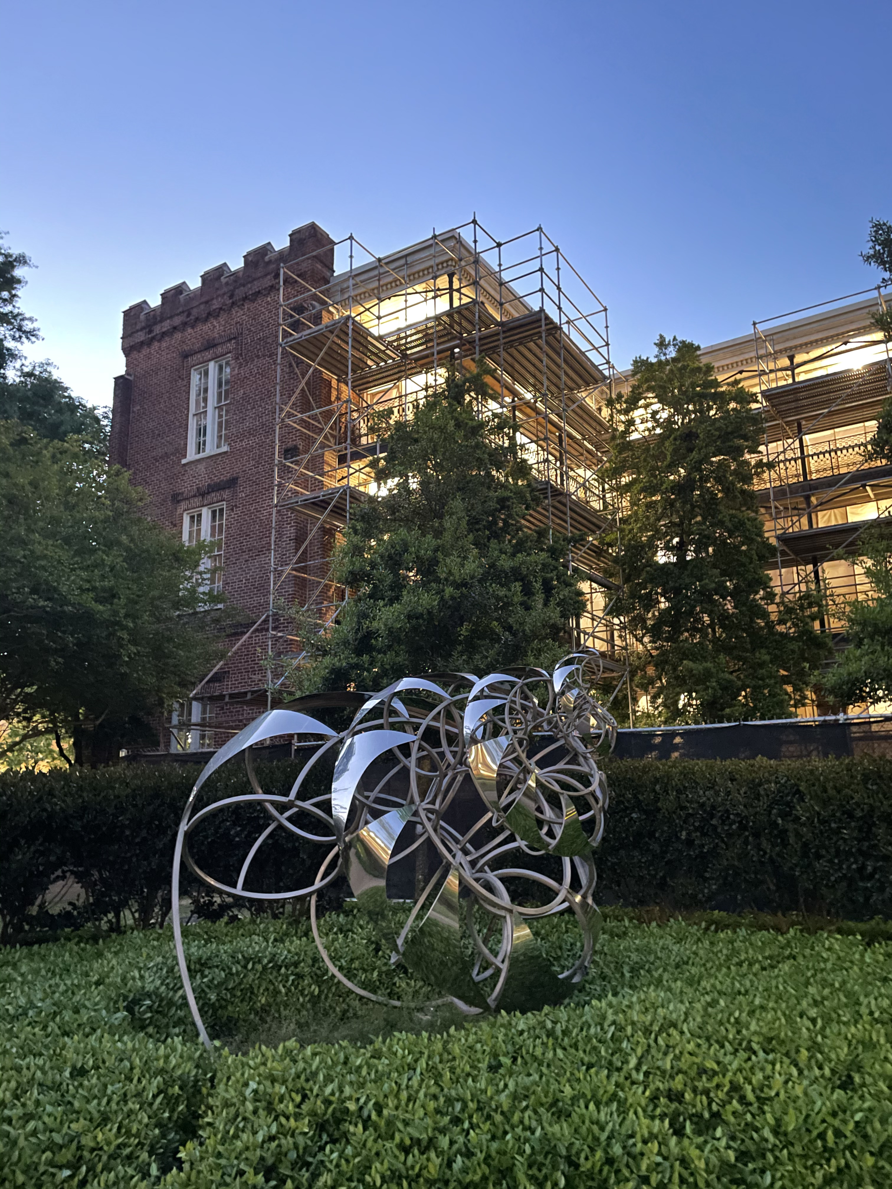 A statue made of thin pieces of shiny metal wrapped up in a spiral. The statue is in a court yard of a brick building with shrubbery all around.
