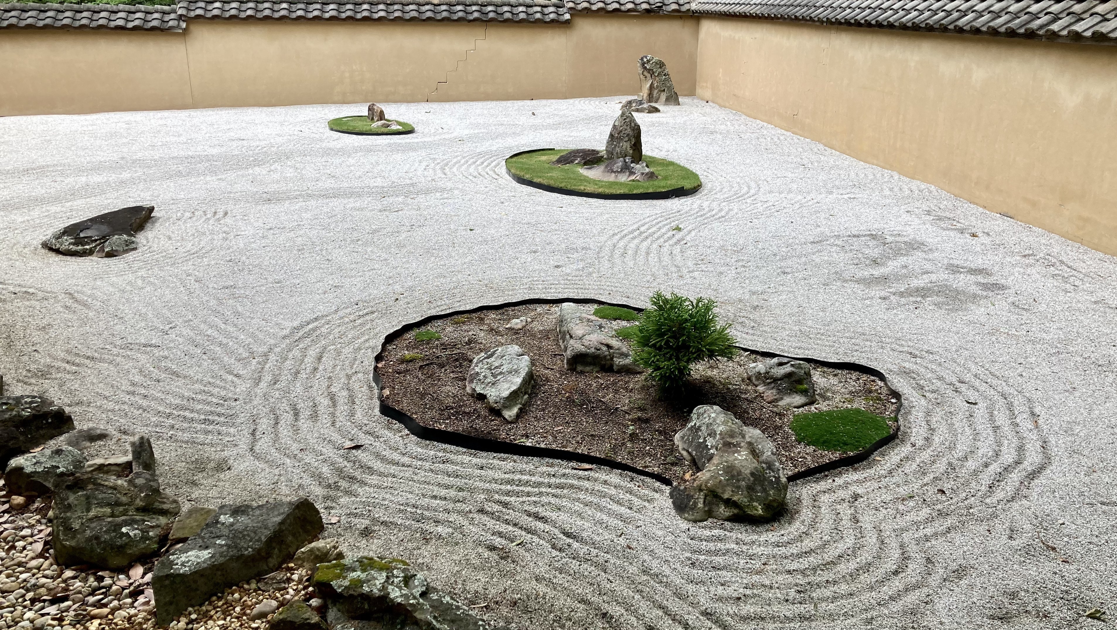 A Japanese rock garden with circular ripples coming from pockets of grass and plants. A wall borders the scene.
