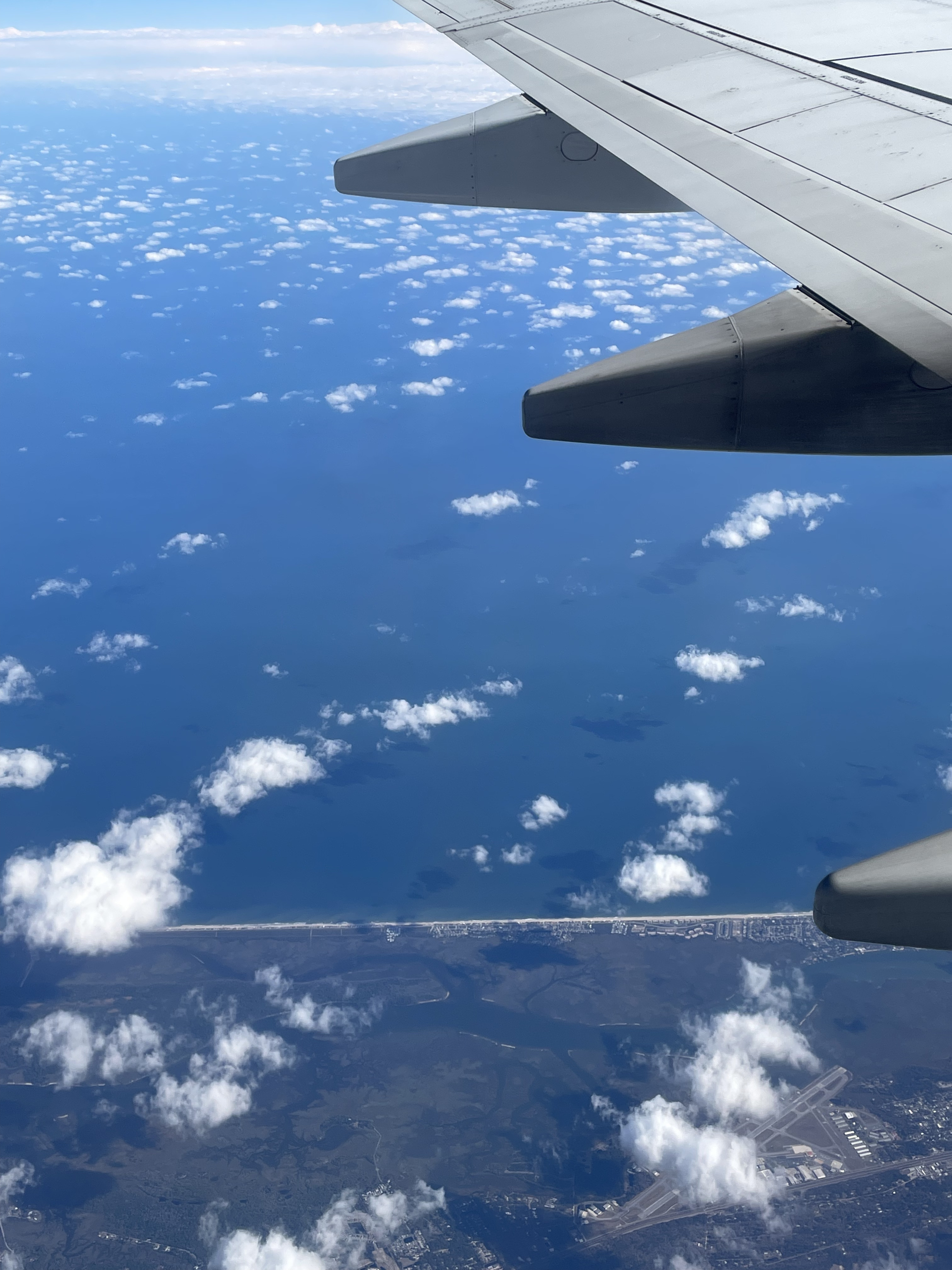 The view outside a plane window. You can see a royal blue ocean and sparse, fluffy clouds near the surface.