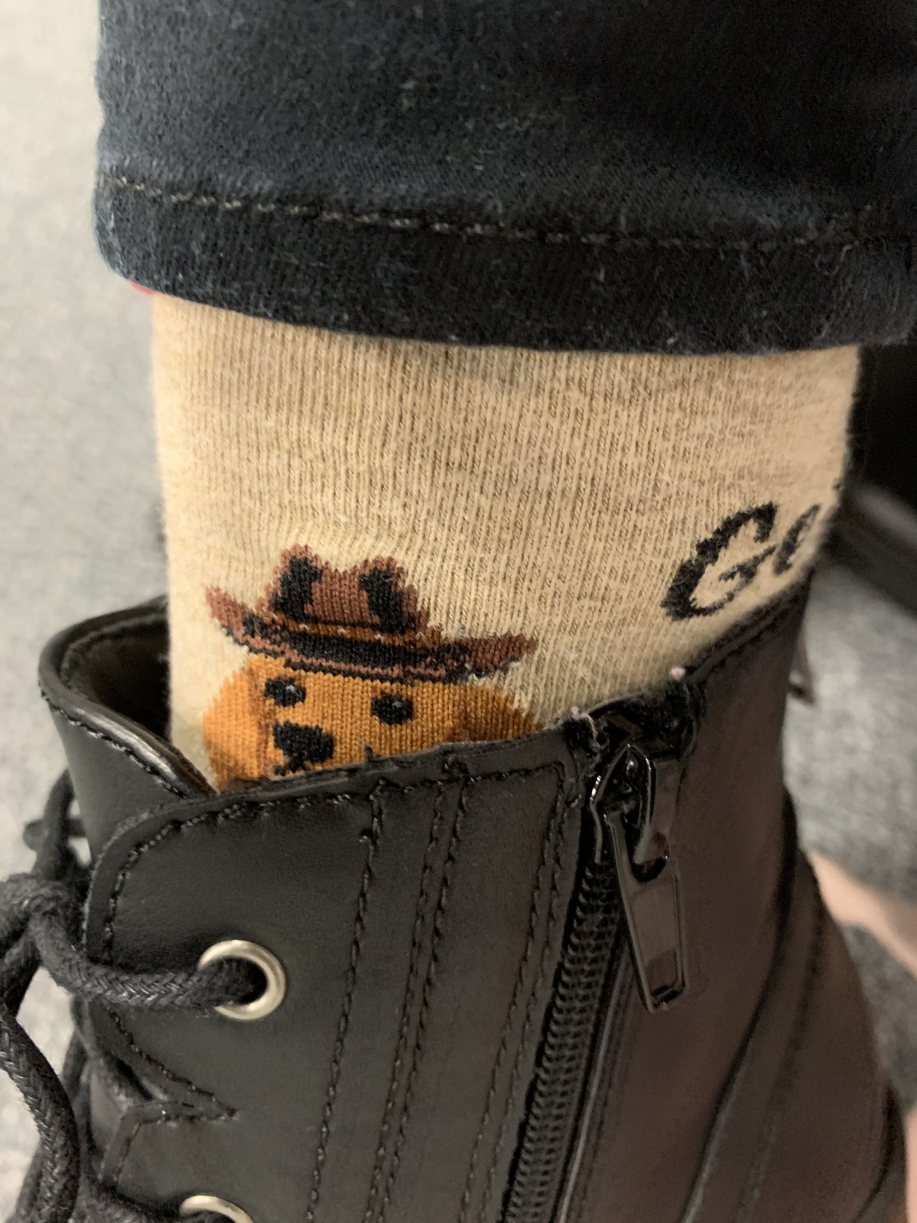 A small section of sock between a pant leg and the top of a black combat boot. The sock has a weiner dog on it that is peeking over the top of the boot. The weiner dog has a cowboy hat.